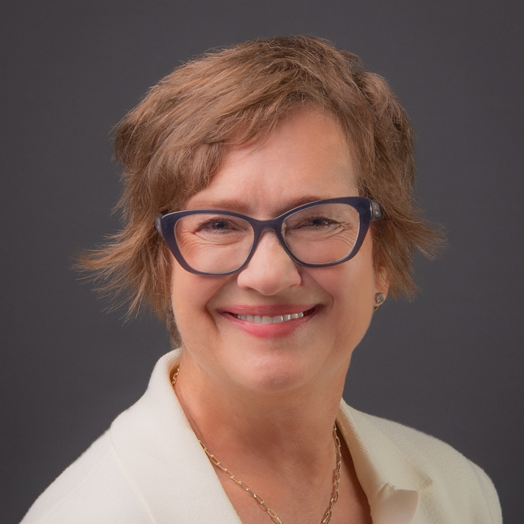 Jackie Hummelbrunner is wearing a cream blazer and dark blue rimmed thicker glasses. She has short chin-length light brown hair and is light skinned.