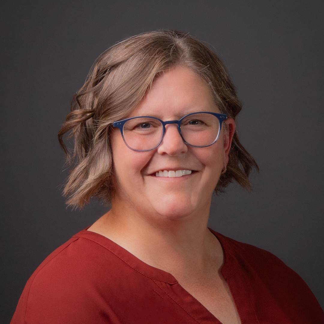 Michelle Gaudet smiles for the camera, she is wearing a red blouse and dark blue rimmed glasses. She has curled her light brown hair.