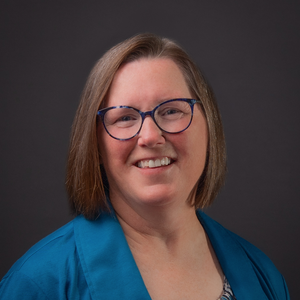 Tammy Hanstead is wearing a bright blue blazer and dark blue rimmed glasses. She has light brown hair that falls just below her chin. She is fair skinned.