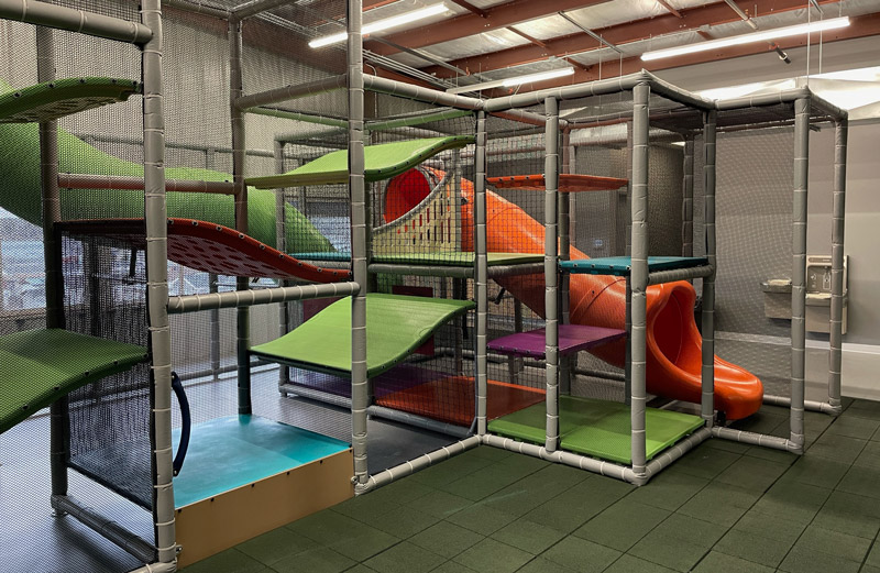 The image shows a giant indoor play area in bright orange, green, purple, blue and yellow. There is a tube slide that's connected to an upper deck that children can access through bright green platforms. The whole structure is surrounded by netting.