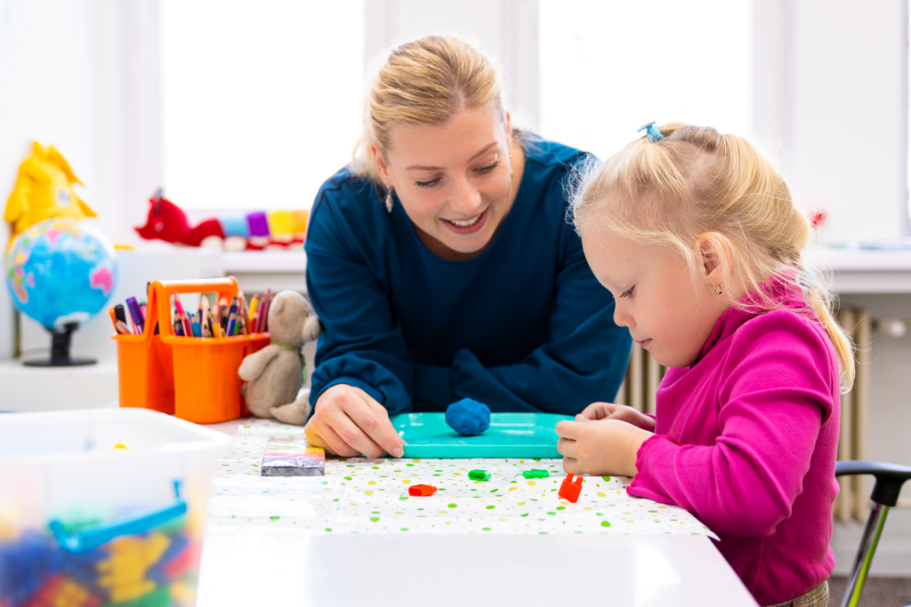 An older white woman with blonde hair in a ponytail and a blue shirt looks at a little girl as she plays with blue playdough. The little girl is also white with very light blonde hair done in a braid, she is smiling whole she works with the playdough.