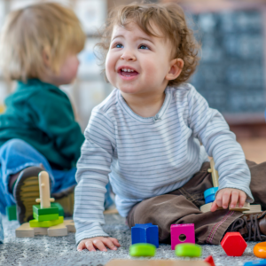 A young child with light skin, blue eyes and dark brown curly hair smiles while looking up. They are playing with brightly coloured wooden blocks on a carpet.