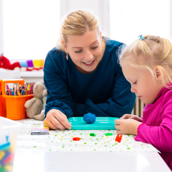 An older white woman with blonde hair in a ponytail and a blue shirt looks at a little girl as she plays with blue playdough. The little girl is also white with very light blonde hair done in a braid, she is smiling whole she works with the playdough.